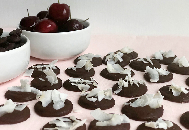 Flat chocolate topped with coconut shavings in front of a bowl of chocolate chips and a bowl of cherries.