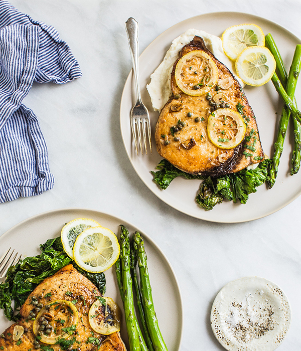 Two plates of swordfish topped with lemon over lettuce and mashed potatoes with a side of asparagus.