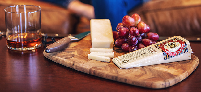 Glass of whiskey next to a cutting board with a white cheese and red grapes.