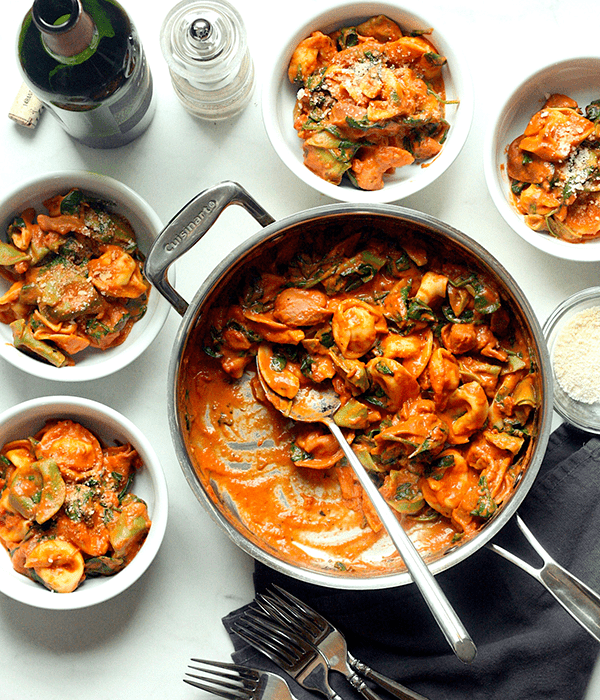 A Pan Full of Tortelloni with Tomato Spinach Cream Sauce Beside Small Serving Bowls