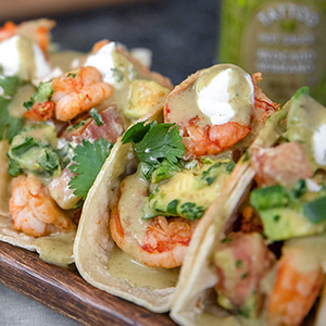 Tacos in a row with shrimp, avocado, green salsa, sour cream, and cilantro.