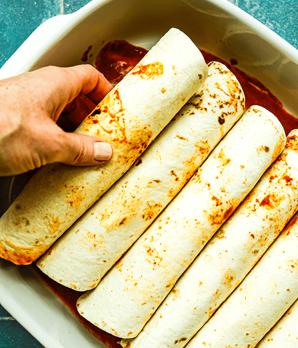 Rolled Enchiladas Being Placed into a Baking Dish with Sauce