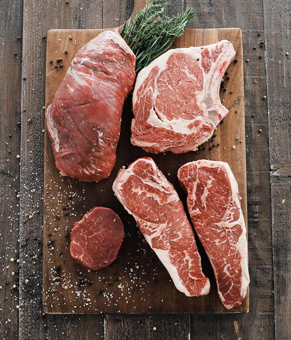 Various cuts of raw steak on a cutting board with salt, pepper, and herbs.