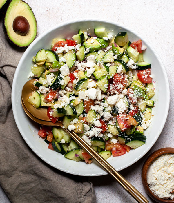 Cucumber salad with tomato and feta in a serving bowl with a gold spoon.