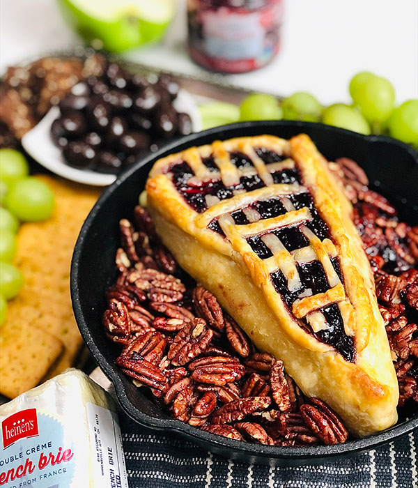 Skillet with brie baked wrapped in a pie crust with cherry pie filling surrounded by roasted pecans.