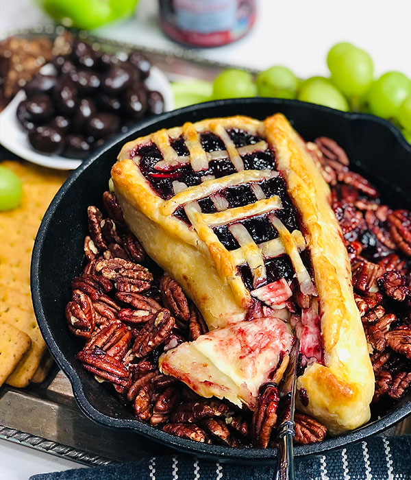 Skillet with brie baked wrapped in a pie crust with cherry pie filling surrounded by roasted pecans.