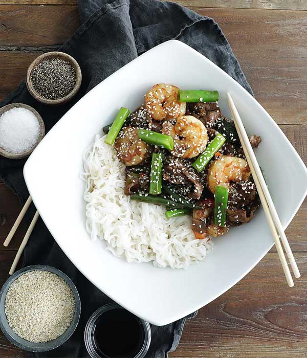 Bowl with rice noodles, shaved steak, shrimp and chopped asparagus topped with sesame seeds, surrounded by small bowls of spices and soy sauce.