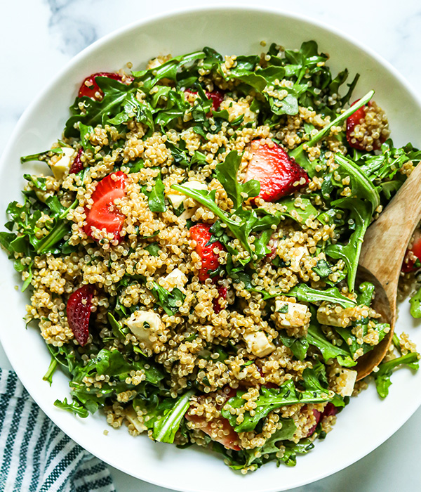 Quinoa salad with strawberries and spring mix in a serving bowl with a wooden serving spoon. 