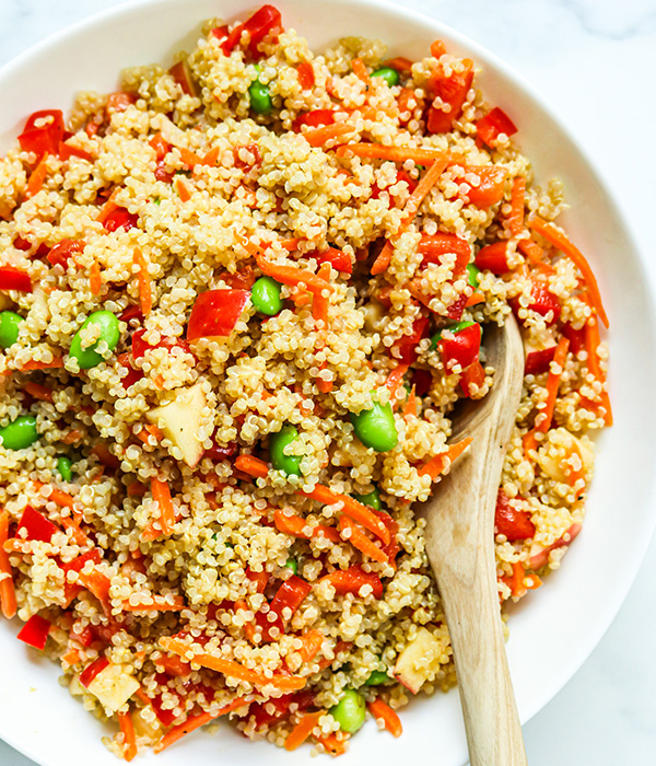 Quinoa salad with tomatoes, carrot sticks, red bell pepper and edamame in a bowl with a wooden serving spoon. 