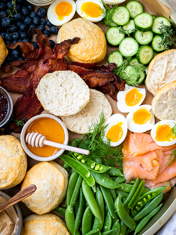 Brunch board with bacon, English muffins, biscuits, salmon, cucumber, hard boiled eggs, blueberries, honey, and snap peas.