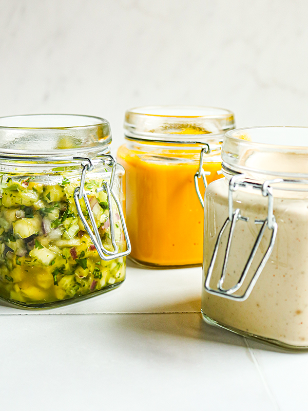 Three condiment jars filled with spicy white BBQ sauce; pineapple, cucumber & red onion relish; and carrot ginger sauce.