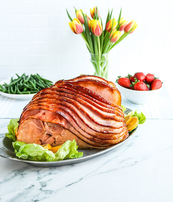 Heinen's Spiral Sliced Ham on a Platter with Fresh Greens. A Heinen's Butcher Shop Box and a Vase of Tulips are in the Background