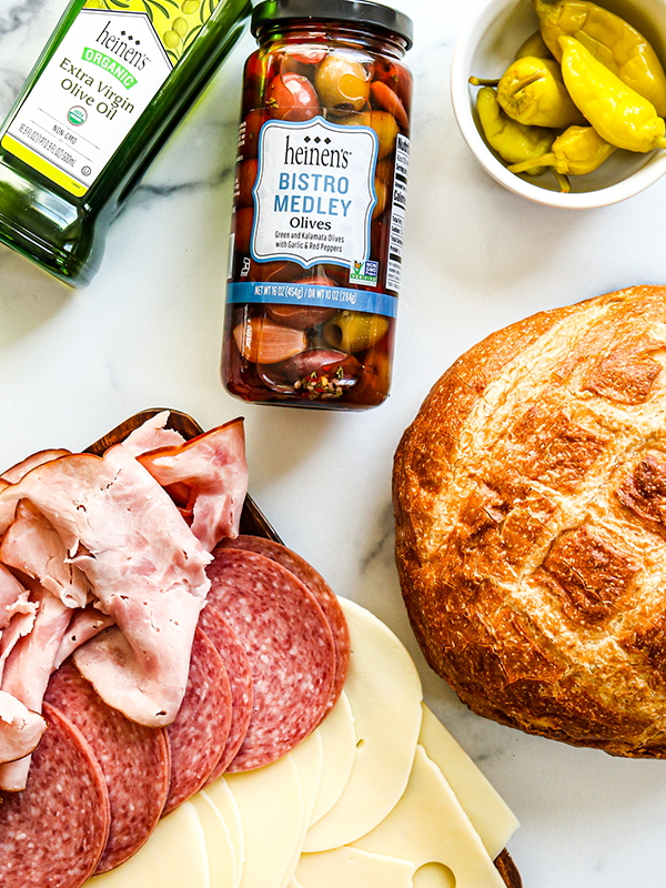 Muffuletta Sandwich ingredients including olives, olive oil, ciabatta bread, pepperoncini, and slice deli meats.