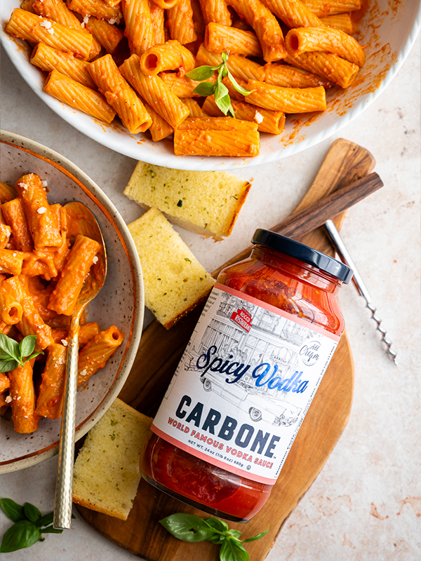 Rigatoni pasta with marinara sauce served in a bowl with a side of garlic bread and placed next to a container of Carbone Spicy Vodka sauce.