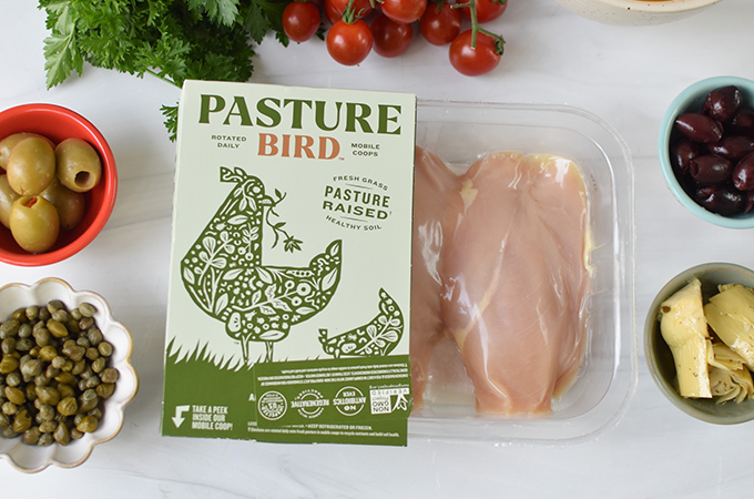 Package of Pasture Bird raw chicken breasts next to olives, capers, and artichoke hearts.