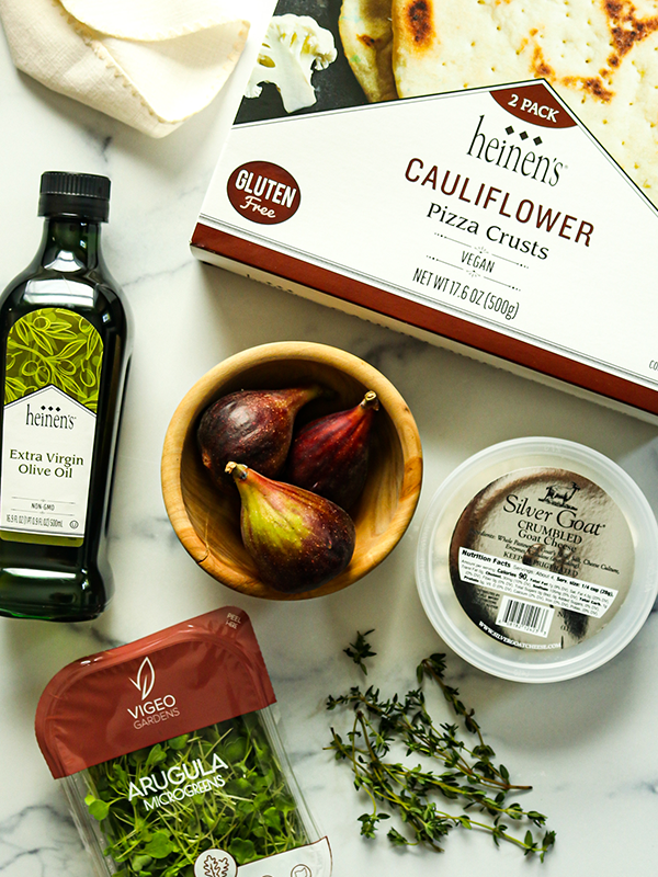 Spread of ingredients including Heinen's cauliflower pizza crusts, olive oil, arugula, goat cheese, and figs.