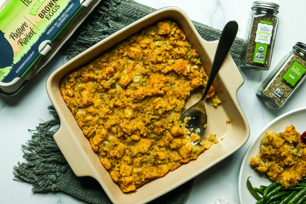 Cornbread dressing in a baking dish next to thyme, sage, and a container of brown eggs.