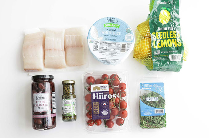 Raw Hakibut Fillets on a White Surface with Heinen's Cooked Rice, a Bag of Lemons, A Jar of Olives, A Jar of Capers, Cherry Tomatoes and Fresh Oregano