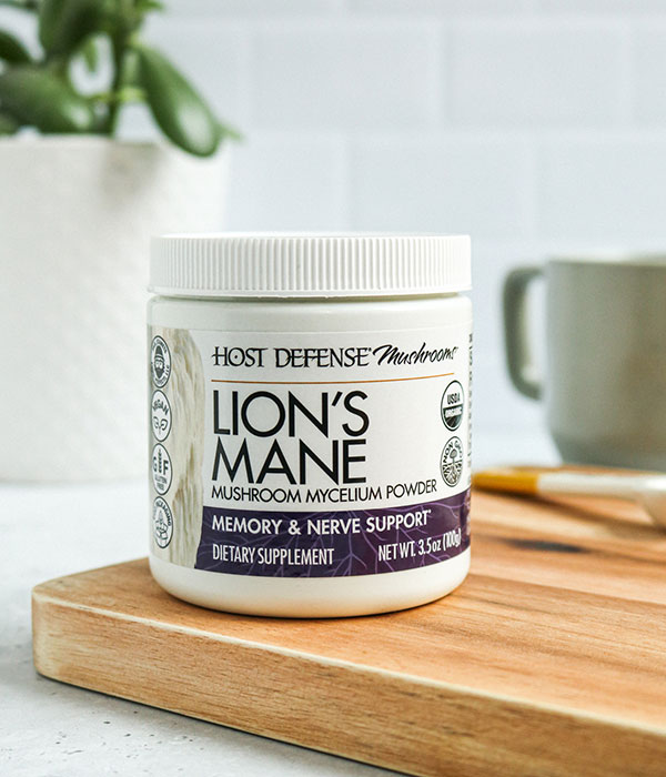 Host Defense Lion's Mane Mushroom Powder in its packaging on a on a wood cutting board with a subway tile background, cup of coffee and a plant.