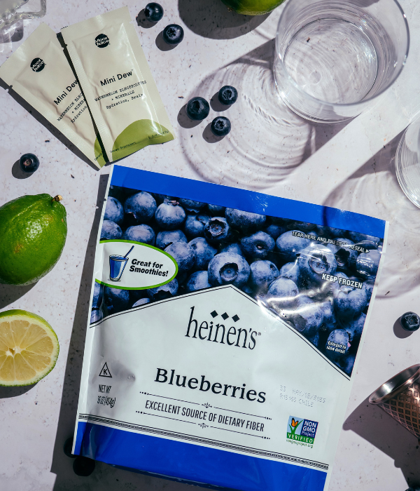 Bag of Heinen's Frozen Blueberries, Moon Juice Mini Dew Watermelon powder packets, and slices of limes next to empty glass.