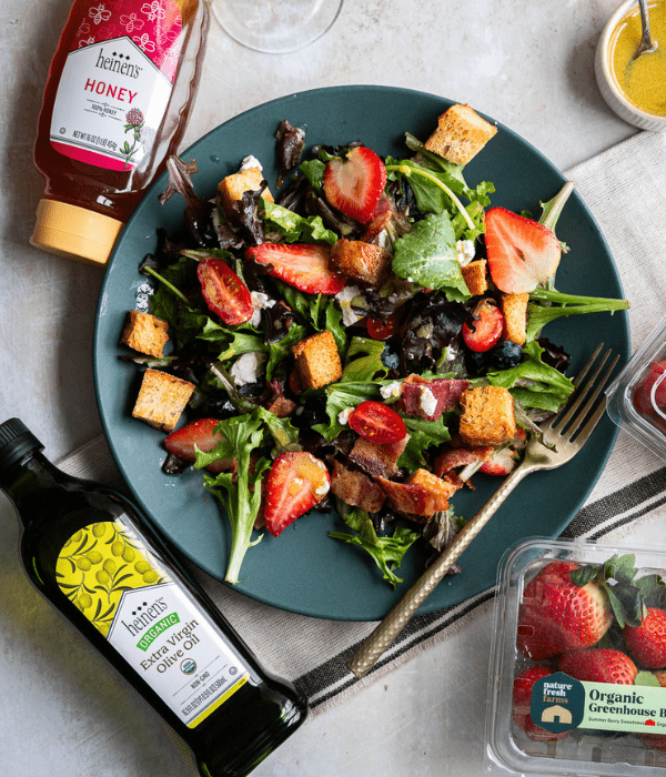 Bacon and Berry Salad in bowl surrounded by Heinen's Extra Virgin Olive Oil, Heinen's Honey, Fresh strawberries.