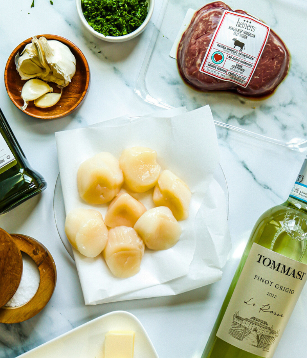 Sea Scallops, Heinen's CARE certified beef tenderloin filet mignons, Garlic, Olivie Oil, and White wine all layed out on white marble table.