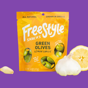 A Package of Heinen's Freestyle Snacking Olives on a Dark Purple Background. Lemon Slices and a Garlic Clove Sit Beside the Package.