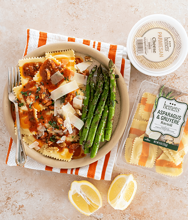 A Bowl of Asparagus and Gruyere Ravioli on an Orange Striped Napkin with a Tomato Butter Sauce and Parmesan Cheese Beside a Pile of Sauteed Asparagus and a Package of Heinen's Asparagus and Gruyere Ravioli, a Package of Heinen's Parmesan Cheese and Fresh Sliced Lemon