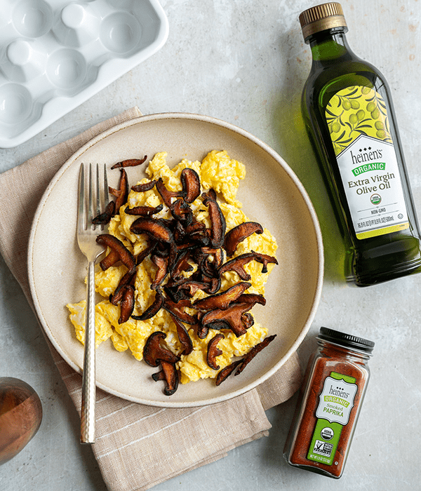 Scrambled Eggs and Shiitake Bacon Alternative in a Bowl with a Fork, a Bottle of Heinen's Extra VIrgin Olive Oil, and a Jar of Heinen's Smoked Paprika