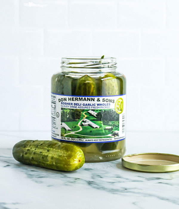 An Open Container of Don Hermann Whole Dill Pickles with a Whole Dill Pickle Sitting Beside the Jar