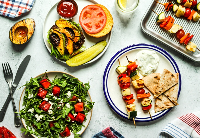 Plates of food including an avocado chicken sandwich, watermelon arugula salad, and halloumi and vegetables with pita bread and tzatziki sauce.