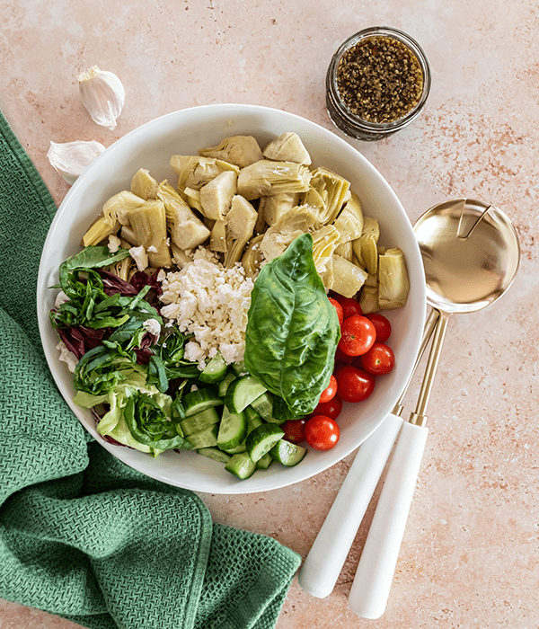 Marinated Artichoke Salad Ingredients in a Serving Bowl with Gold and White Serving Spoons, a Green Cloth Napkin and a Glass Jar of Balsamic Dressing