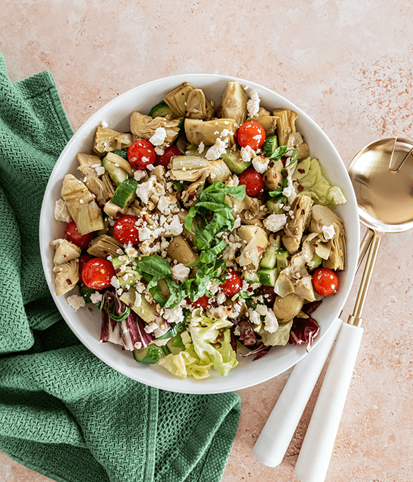 Marinated Artichoke Salad in a Serving Bowl with Gold and White Serving Spoons, a Green Cloth Napkin and a Glass Jar of Balsamic Dressing