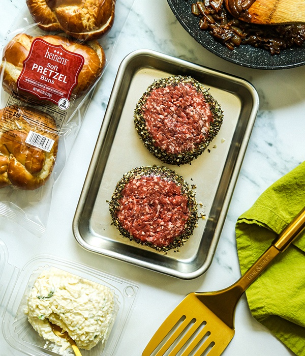 Two Heinen's Gourmet Burger patties on a tray beside a package of pretzel buns and a container of Jarlsberg cheese spread.
