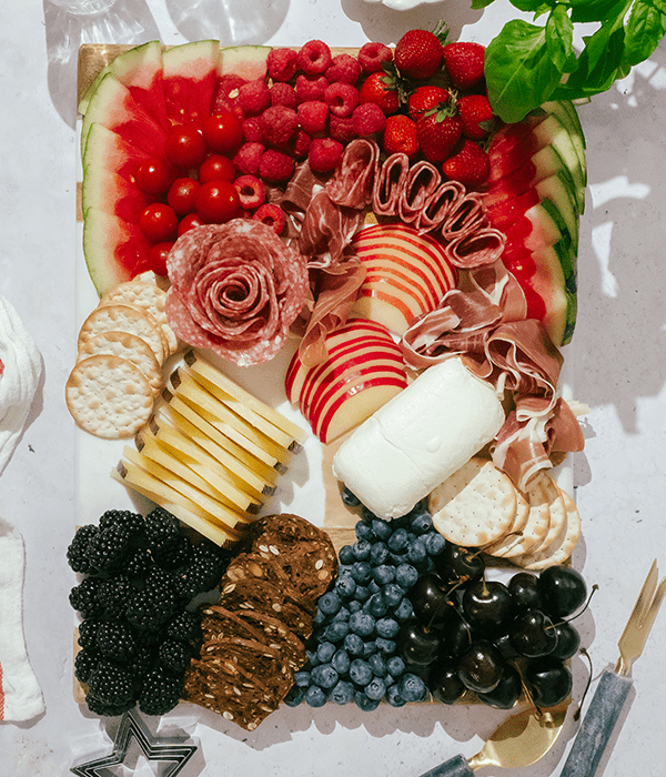 A Partially Assembled Charcuterie Board with Sliced Cheeses, A Whole Goat Cheese Log, Sliced Fruit, Rolled Charcuterie Meats, Red Berries, Blackberries, Blueberries and Cherries.