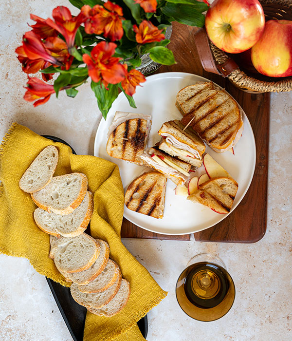 Turkey, Apple, and Cheddar Panini sandwich on a plate with a basket of apples and sliced bread.