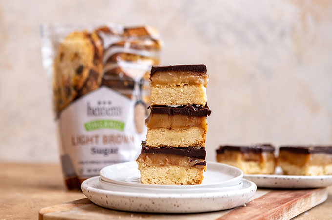 Plate with three stacked pieces of millionaire's shortbread: shortbread topped with caramel and chocolate and a bag of Heinen's brown sugar