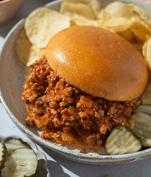 A Sloppy Joe Sandwich on a Plate Beside a pile of Kettle Chips and Dill Pickle Slices