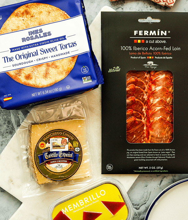 A Rectangular Stone Charcuterie Board with a Package of Sourdough Torta Crackers, a Block of Manchego Cheese, a Package of Iberico Ham, and a Package of Membrillo Quince Paste