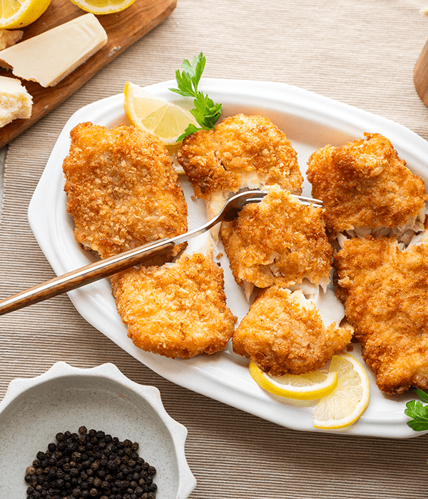 Three Parmesan Crusted Cod Fillets in a White Serving Platter with a Fork and Lemon Wedges