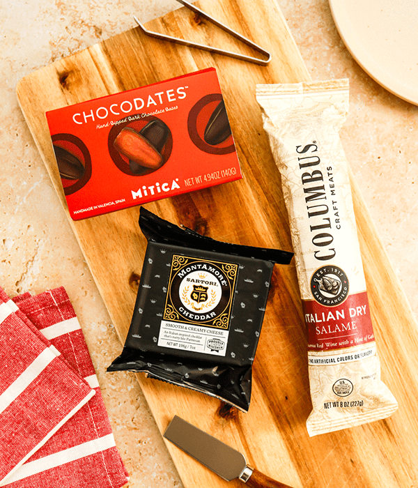 A Rectangular Wood Charcuterie Board with a Package of Wisconsin Cheddar Cheese, a Chubb of Salami, and a Box of Chocolate Dates