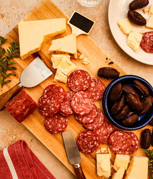 A Rectangular Wood Charcuterie Board with a Block of Sliced Wisconsin Cheddar Cheese, a Pile of Sliced Salami, and a Bowl of Chocolate Dates