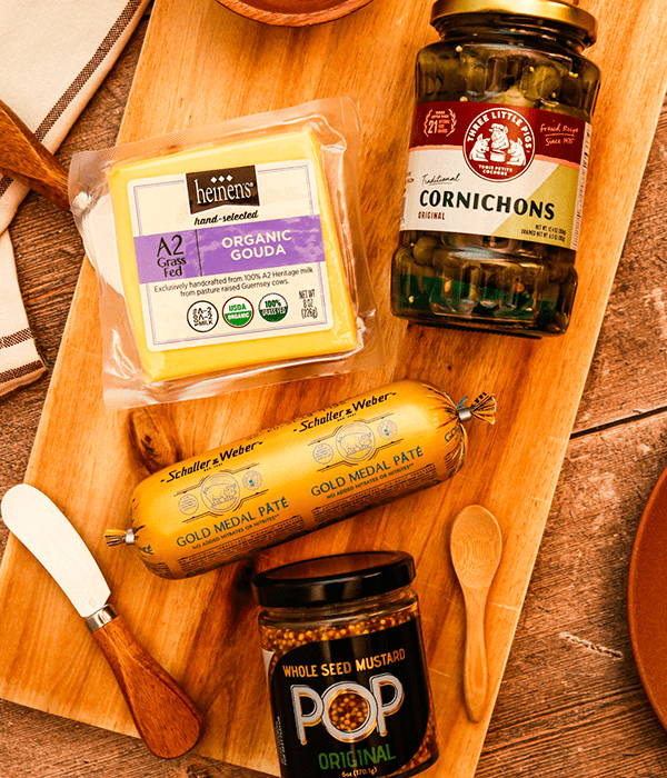 A Rectangular Wooden Charcuterie Board with a Chub of Pork Pâté, a Block of Gouda Cheese, a Container of Cornichons, and a Container of Grainy Mustard