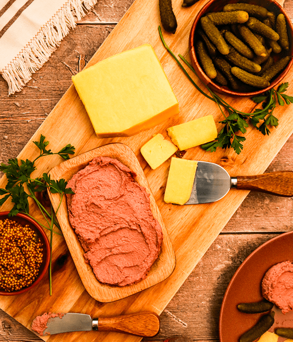 A Rectangular Wooden Charcuterie Board with Sliced Gouda Cheese, a Bowl of Cornichons, a Plate with Pork Pâté, and a Bowl with Grainy Mustard