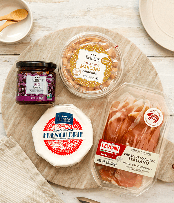 A Charcuterie Board with a Container of Fig Spread, a Wheel of French Brie, a Package of Prosciutto, and a Package of Marcona Almonds