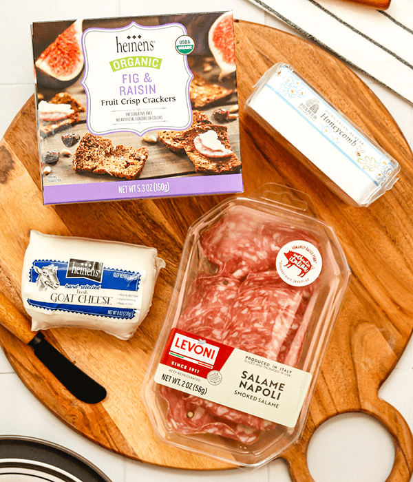 A Round Wooden Charcuterie Board with a box of fig and raisin fruit crisp crackers, A Block of Gaot Cheese, a Package of Salami, and a Honeycomb Package