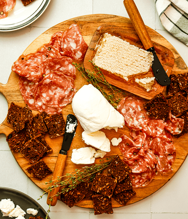 A Round Wooden Charcuterie Board with Sliced Goat Cheese, Piles of Salami, a Hungarian Honeycomb, and Fig and Fruit and Raisin Crackers