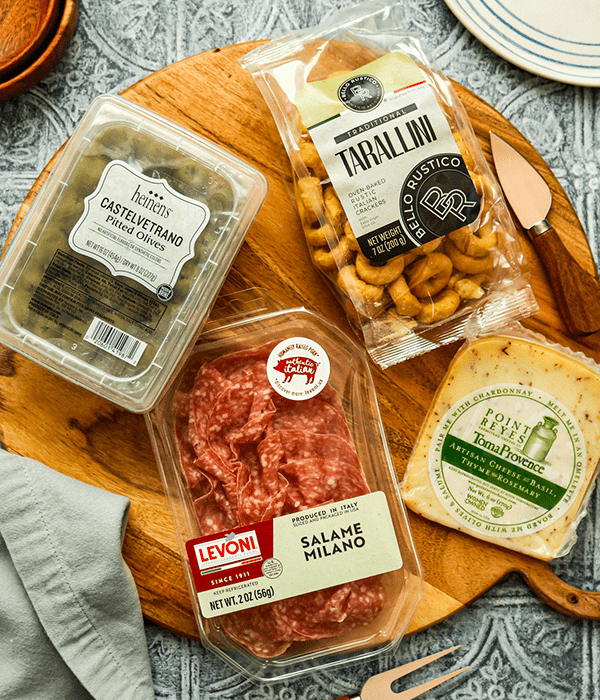 A Charcuterie Board with a Package of Castelvetrano Olives, a Block of Point Reyes Cheese, a Package of Salami, and a Bag of Knot Crackers