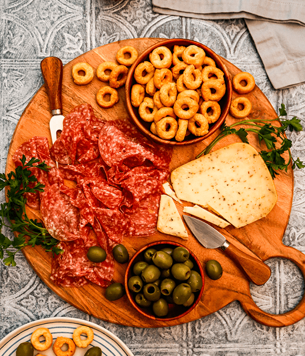 A Charcuterie Board with a Block of Sliced Cheese, a Bowl of Castelvetrano Olives, a Bowl of Round Knot Crackers, and a Pile of Salami