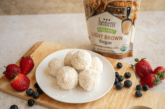 6 No Bake Coconut Snowballs on a wooden paddle with blueberries, strawberries, and Heinen's light brown sugar